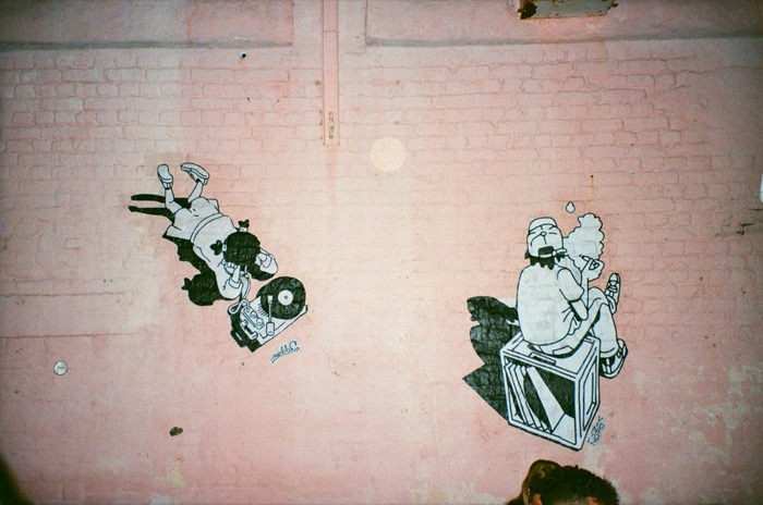 Two wheatpaste DJ characters mid-motion on a pink brick wall, illustration by Sakkie